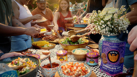 People gather around a table outdoors filled with bowls of salad, corn, salsa, and snacks during a meal.