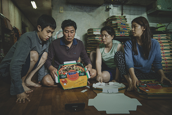 Four people sit on a wooden floor in a dimly lit room, staring intently at an open pizza box.