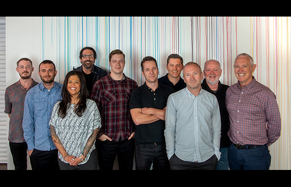 A group of nine men and one woman standing together in a line against a colorful striped wall.