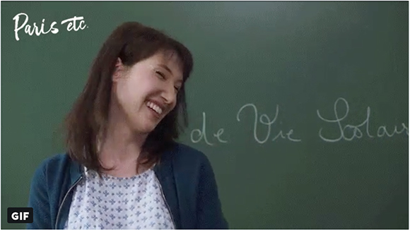 A woman with brown hair laughs while standing in front of a green chalkboard with white writing.