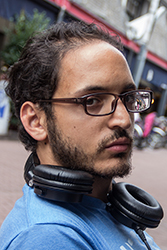 A man with dark hair, glasses, and a beard wearing black headphones around his neck.