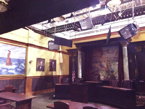 A courtroom with wood paneling, portraits on the wall, and studio lighting rigs hanging from the ceiling.