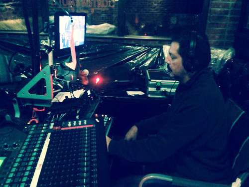 A man sits at a desk with a large mixing console and computer monitor in a dimly lit room.