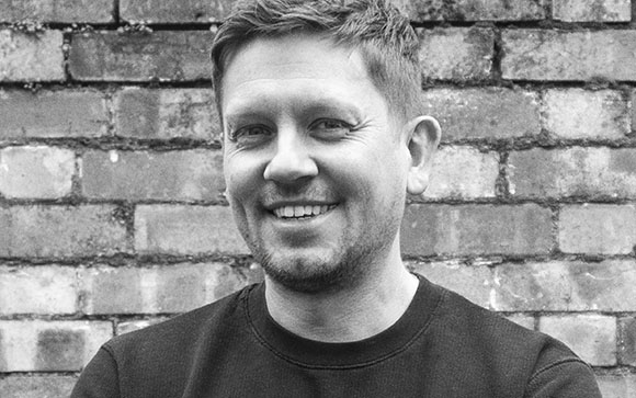 A smiling man in a dark t-shirt posing in front of a brick wall.