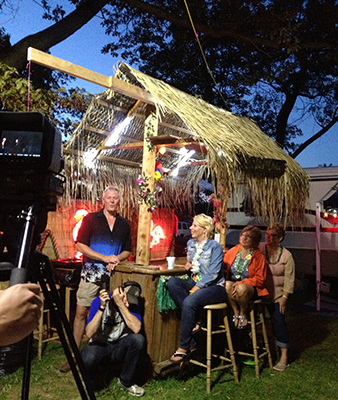 Four people stand and sit near a small wooden hut with a straw roof under trees at dusk.