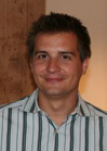 A man with short dark hair smiling while wearing a white and dark vertical striped shirt.