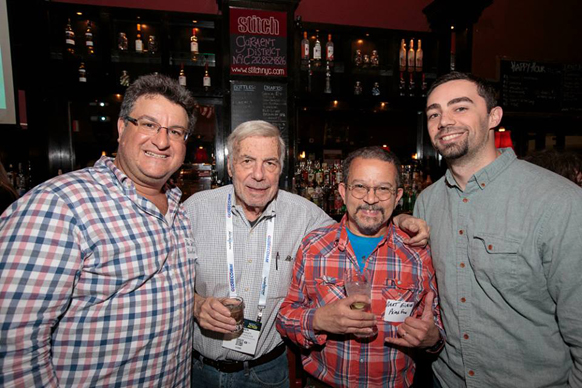 Four men smiling and posing for a photo inside a dimly lit bar with liquor bottles on shelves behind them.