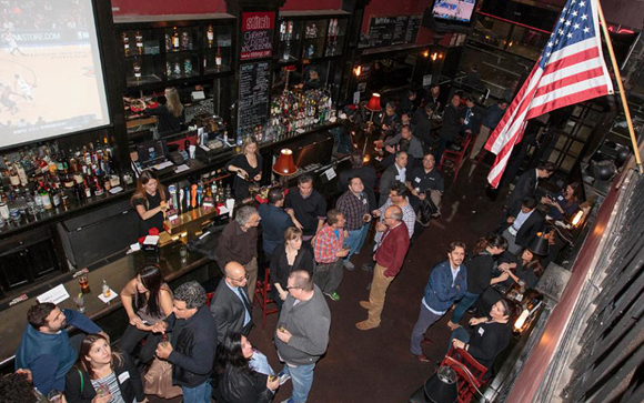 High-angle shot of many people standing and sitting in a dimly lit bar with a large American flag.