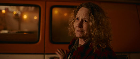 A woman with curly blonde hair and a worried expression stands in front of an orange van with windows.