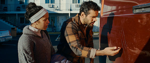 A man with a beard wearing a plaid shirt points to deep scratches on the side of a red van. A woman with a headband and gray