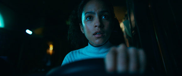 A young woman with wide eyes grips a steering wheel while driving at night, illuminated by streetlights.
