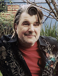 A man with graying hair, a red shirt, and a black shirt with ornate embroidery smiles outdoors.