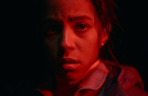 Close-up of a young woman's face, lit by red light, with a somber expression.
