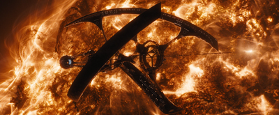 A dark, crescent-shaped spacecraft flying in front of a bright, swirling orange and yellow sun.