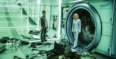 A man and a woman stand in a brightly lit, sci-fi corridor filled with scattered equipment and a large circular hatch.