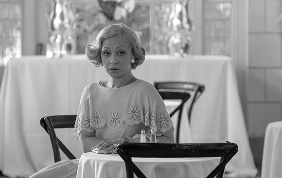 A woman with wavy blonde hair sits at a white-clothed table in a black and white restaurant setting.