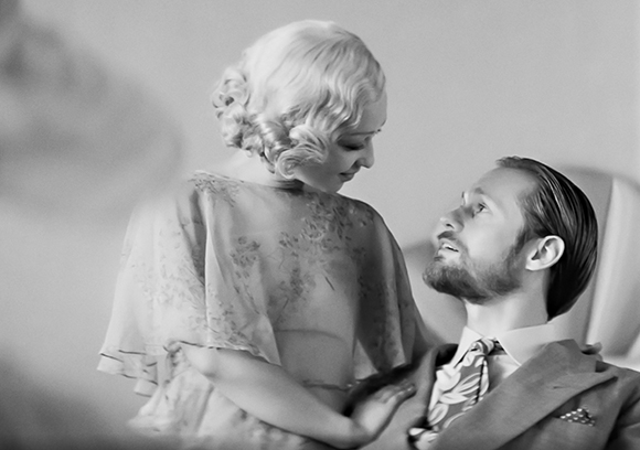 Black and white portrait of a blonde woman in a floral blouse looking down at a bearded man in a suit.