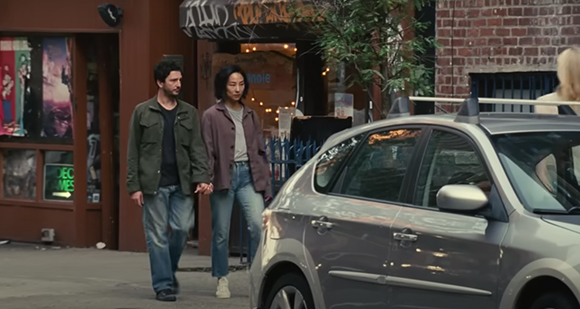 A man in a green jacket and a woman in a purple jacket walk along a sidewalk next to a parked car.