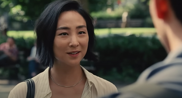 A woman with short black hair smiles while looking at a man in a sunlit park.