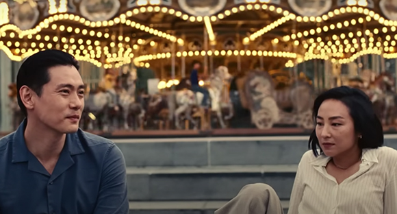 A man and a woman sit in the foreground while a brightly lit carousel glows in the background.