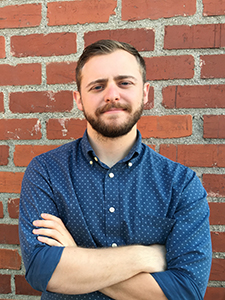 A man with a beard and a blue patterned button-down shirt stands with arms crossed against a red brick wall.