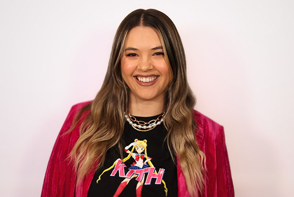 Smiling woman with long blonde hair wearing a black Sailor Moon t-shirt and pink velvet jacket.