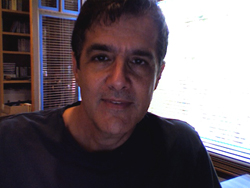A man with dark hair and a dark shirt looking directly at the camera in a room with a window.
