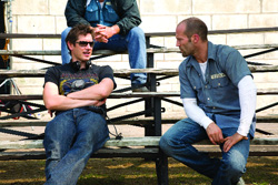 Two men sit on outdoor wooden bleachers, one in a black t-shirt and one in a blue button-down shirt.