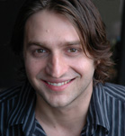 A smiling man with medium-length brown hair wearing a dark striped shirt.