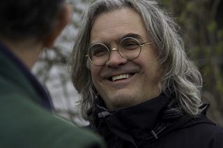 A smiling man with long grey hair and round glasses looks toward another person.