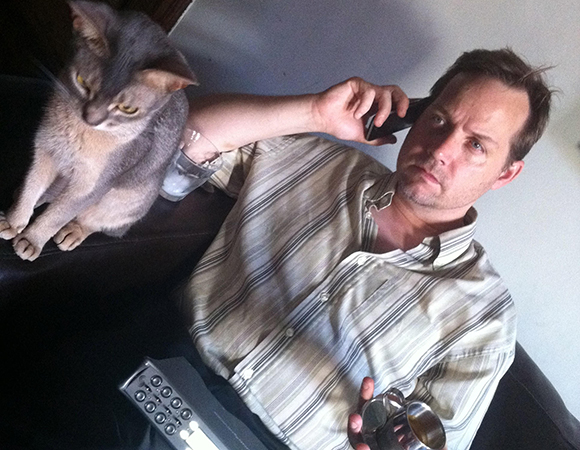 A man in a striped shirt sits on a dark couch, holding a phone to his ear next to a grey cat.
