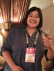 A woman with short dark hair smiles and holds a glass of white wine. She is wearing a blue collared shirt and a lanyard with