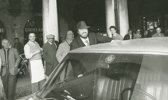 A man in a fedora leans against a car in a crowded nighttime street scene.