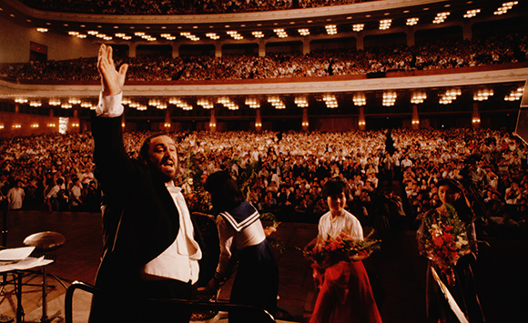 A conductor with his arm raised stands on a stage before a massive, crowded concert hall audience.