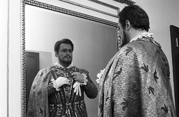 A man in an ornate, patterned cloak and doublet looks at his reflection in a large framed mirror.