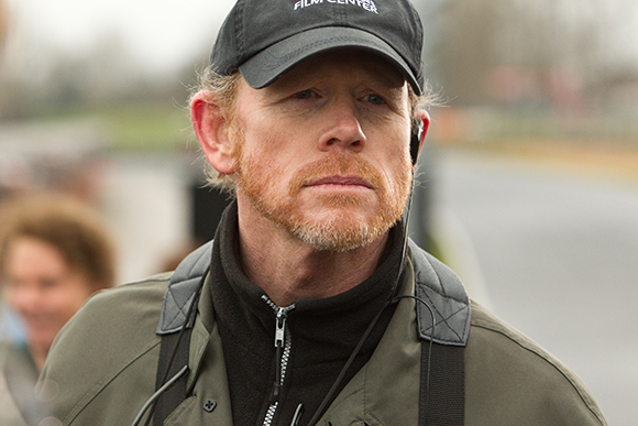 A man with reddish hair and a beard wearing a black cap and an olive green jacket outdoors.