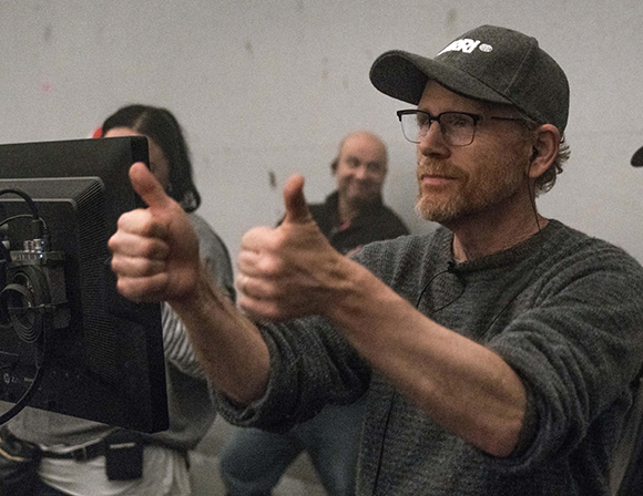 A man wearing a cap and glasses gives two thumbs up toward a computer monitor.
