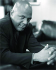 Black and white portrait of a bald man in a dark suit looking toward the camera with hands clasped.