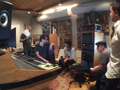 A group of people are gathered in a recording studio, with a soundboard in the foreground and audio equipment in the backgrou