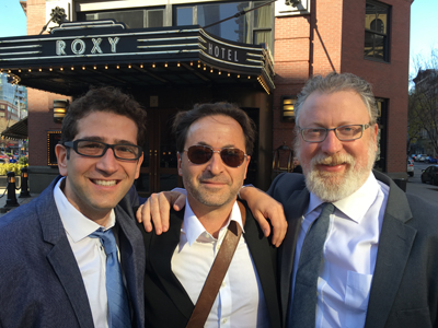 Three men stand in front of the Roxy Hotel. The man on the left wears glasses and a blue tie. The man in the middle wears sun