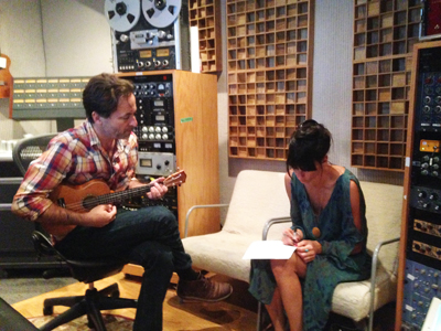 A man plays a ukulele while a woman writes in a notebook in a recording studio with soundproofing panels on the walls.