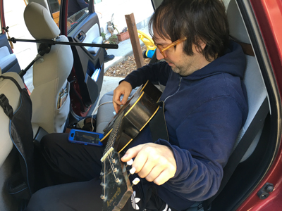 A man with glasses and a blue hoodie sits in the back seat of a car, playing an acoustic guitar. A microphone is mounted on t