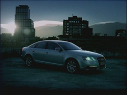 A silver sedan parked on a dark surface with city buildings and mountains in the background at dusk.