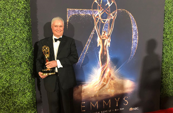 A smiling man in a black tuxedo holds a gold Emmy award in front of a 70th Emmys backdrop.