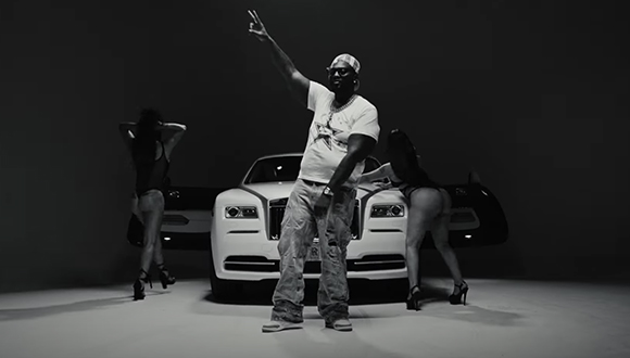 A man in a white t-shirt and baggy jeans poses with a peace sign in front of a white luxury car.