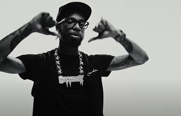 A man wearing a black cap and t-shirt with a large necklace makes hand gestures against a plain background.