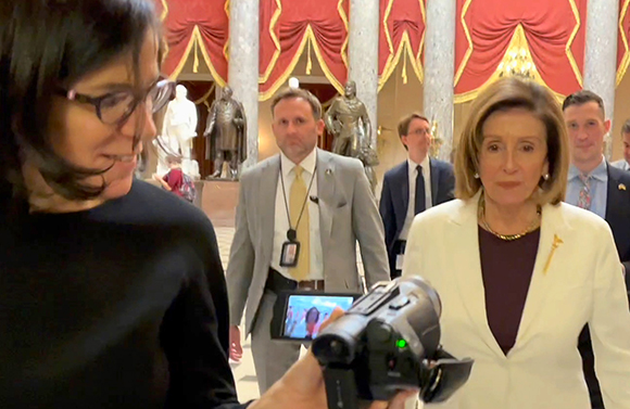 A woman films Nancy Pelosi and several men in a formal hallway with red curtains.