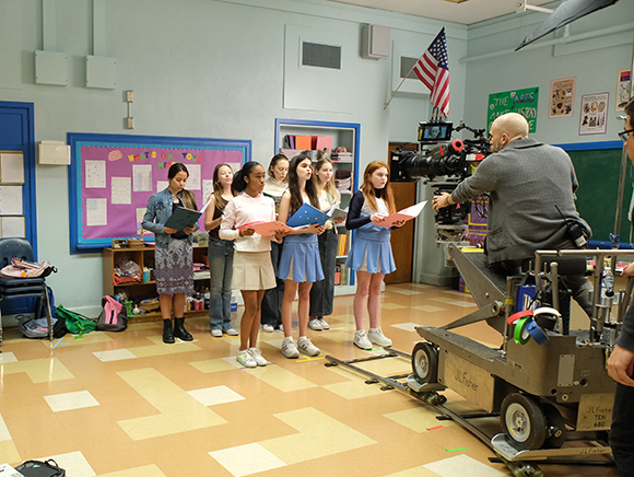 A group of young women stand in a classroom holding scripts while a camera operator films them.