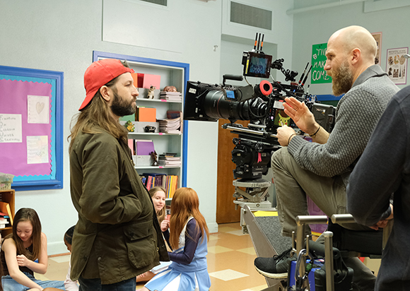 A man behind a large professional cinema camera looks toward another man on a film set.