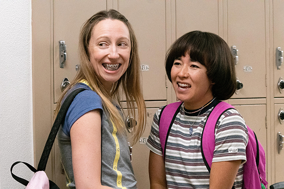 Two smiling students with backpacks standing in front of school lockers.